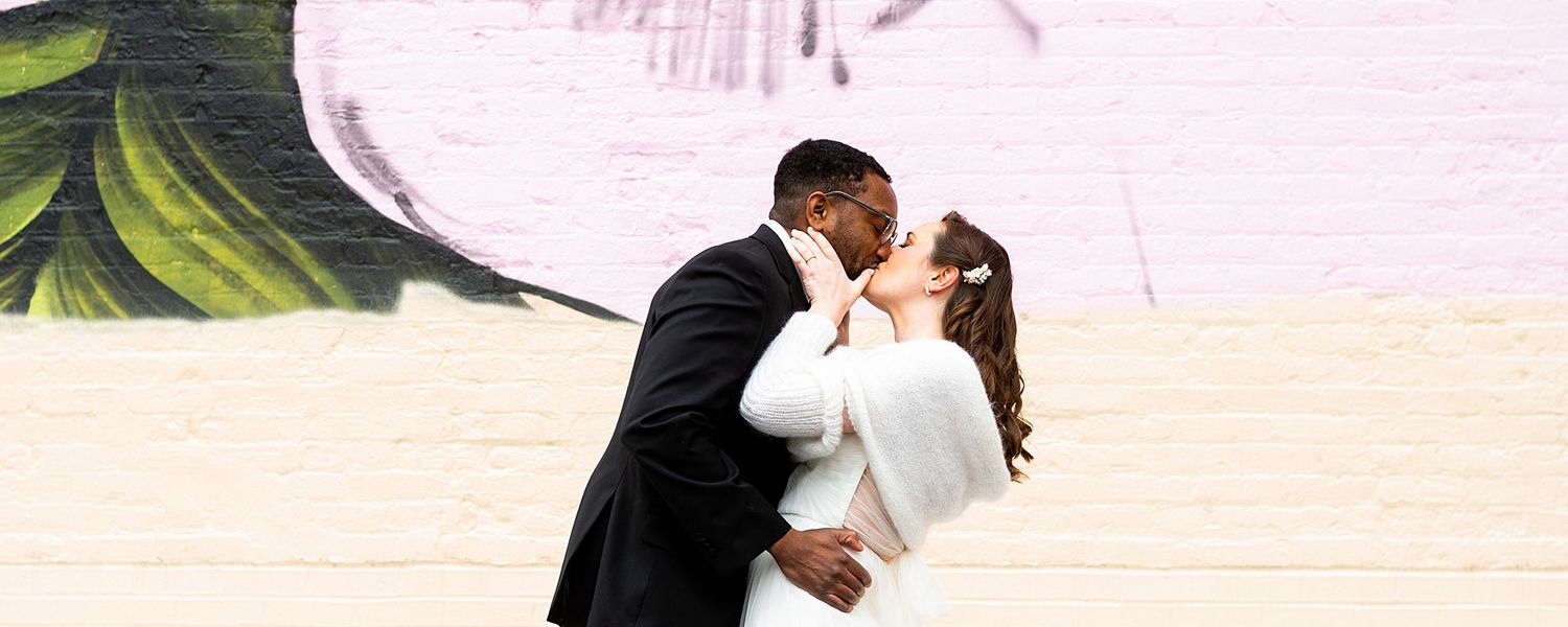 A couple in formal attire share a kiss in front of a light-colored wall with partial green foliage visible.