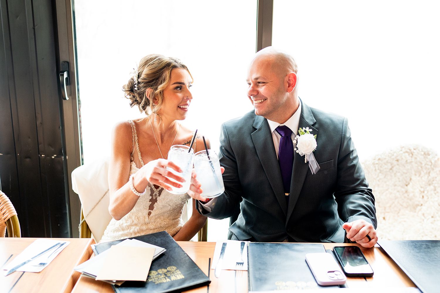 A candid moment shows a bride and groom sharing laughter while signing documents at a desk.