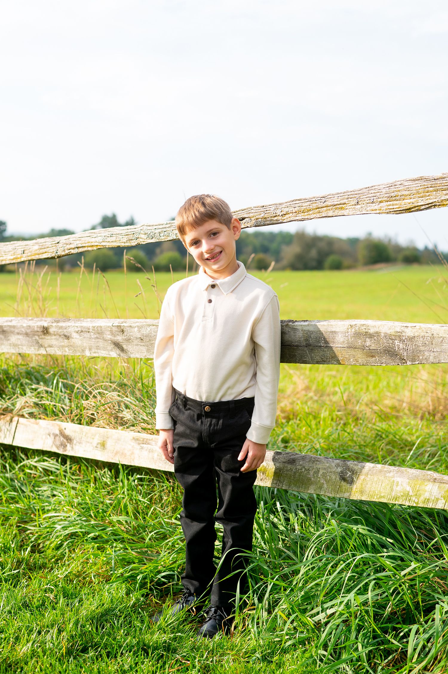 Smiling boy in white shirt and black pants standing by a rustic wooden fence in a green countryside field.