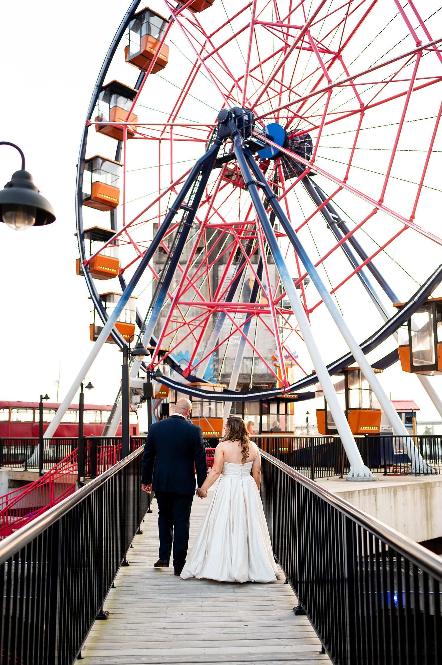 Wedding couple walking along a boardwalk with a large red Ferris wheel structure behind them.