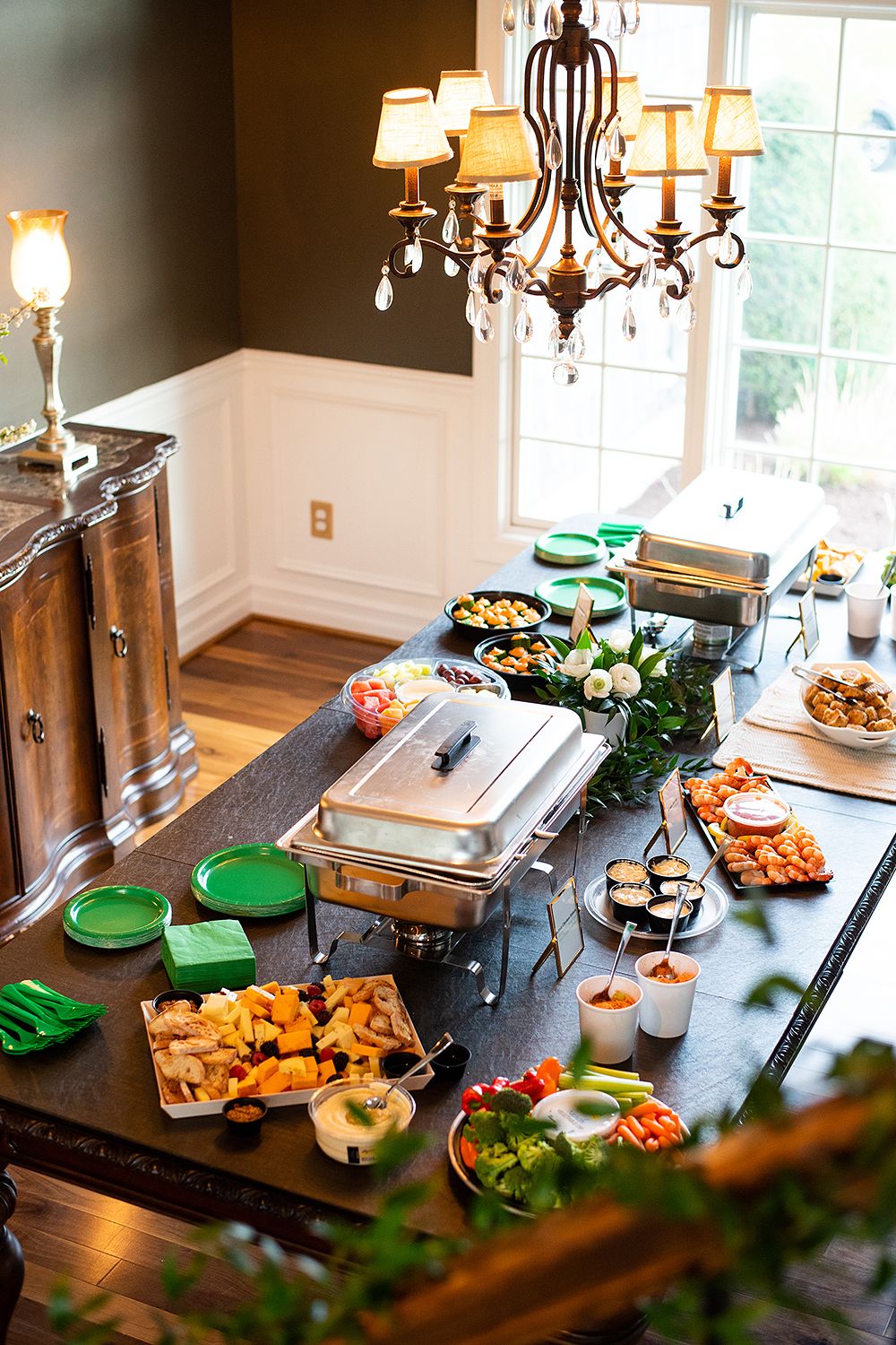 Elegant buffet spread with chafing dishes and appetizers on dark countertop under crystal chandelier in dining room.