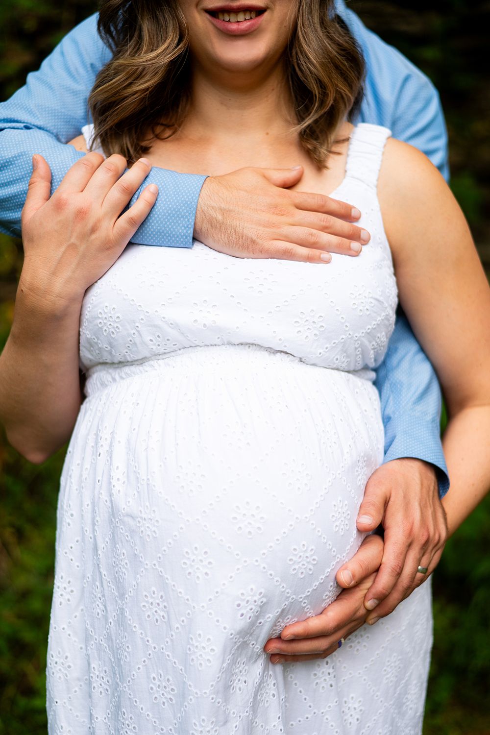A pregnant woman in a white eyelet dress embraces her baby bump while hands gently rest on her stomach.