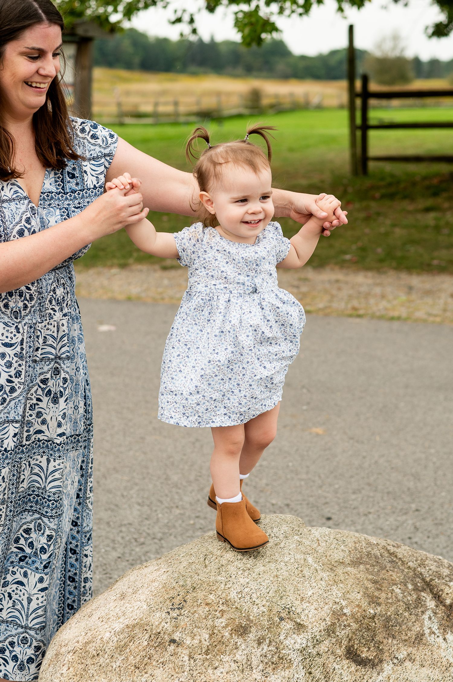 Mother helping smiling toddler girl balance on a rock outdoors, both wearing floral dresses in a park setting.