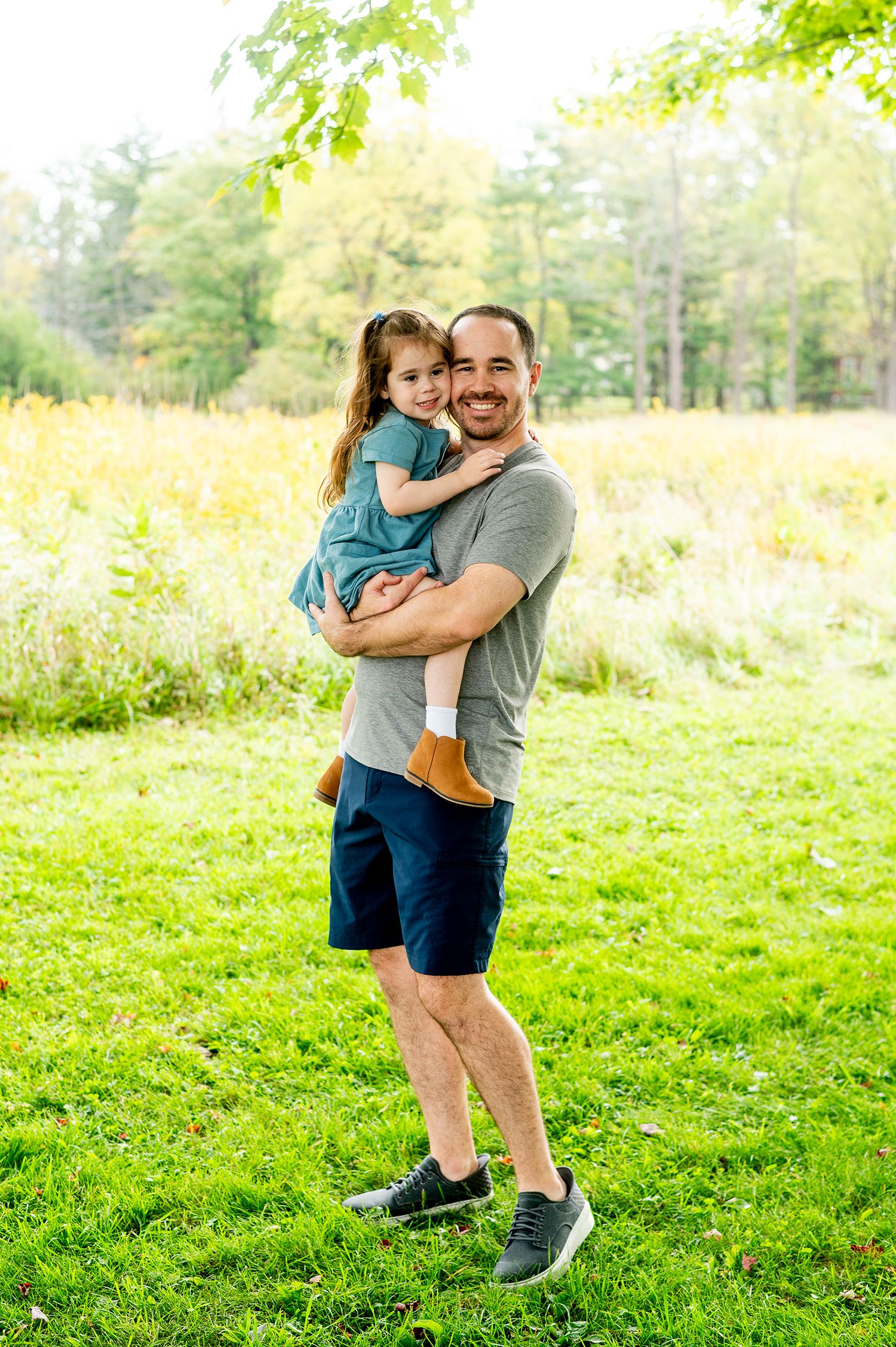 Smiling father holding young daughter in blue dress outdoors on green grass with colorful autumn trees in background.