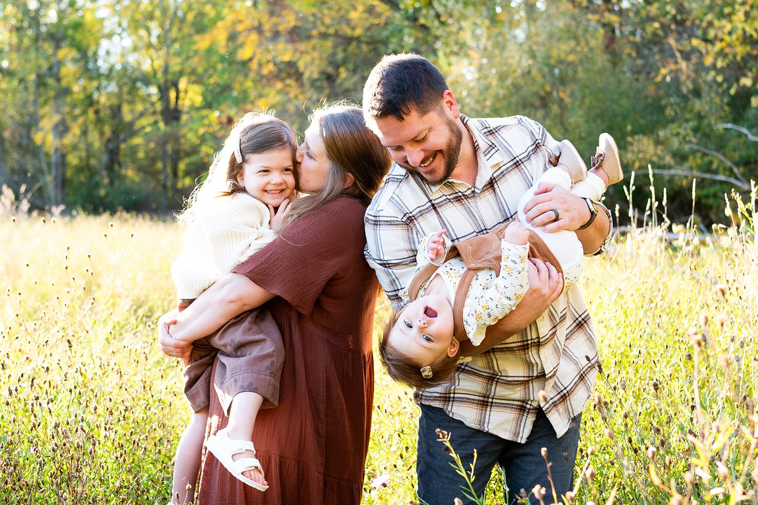 Family of four playing in a sunny field, parents holding laughing children outdoors in autumn.