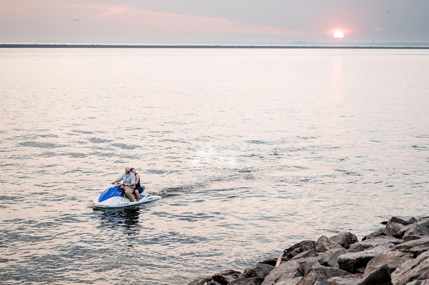 A person rides a blue jet ski on calm waters near rocky shore at sunset with pastel sky.