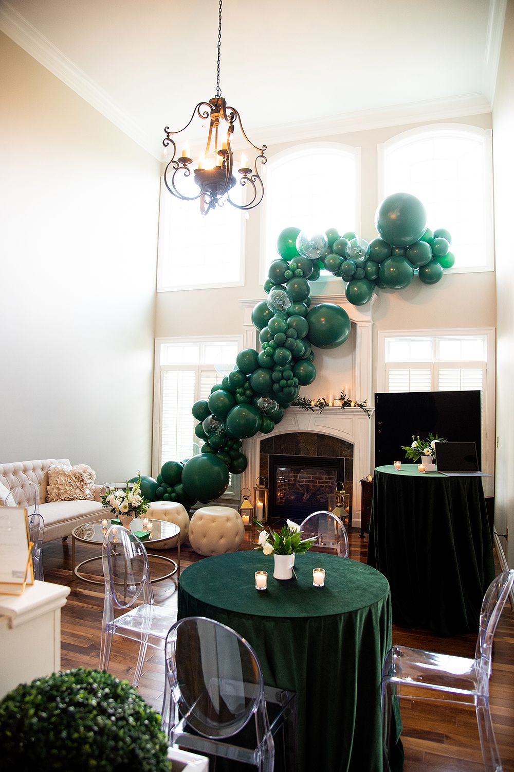 Elegant party setup with emerald green balloon arch, ghost chairs, and chandelier in a bright white room with fireplace.