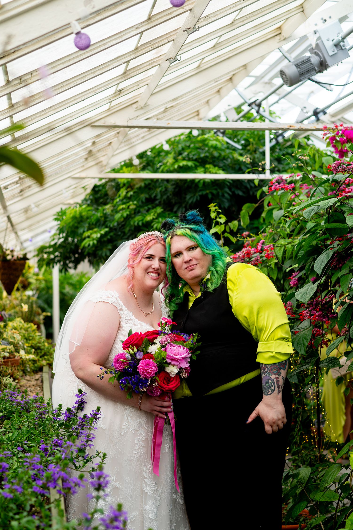Two brides pose in a greenhouse, one in white lace gown with colorful bouquet, other in black and yellow outfit with green hair.