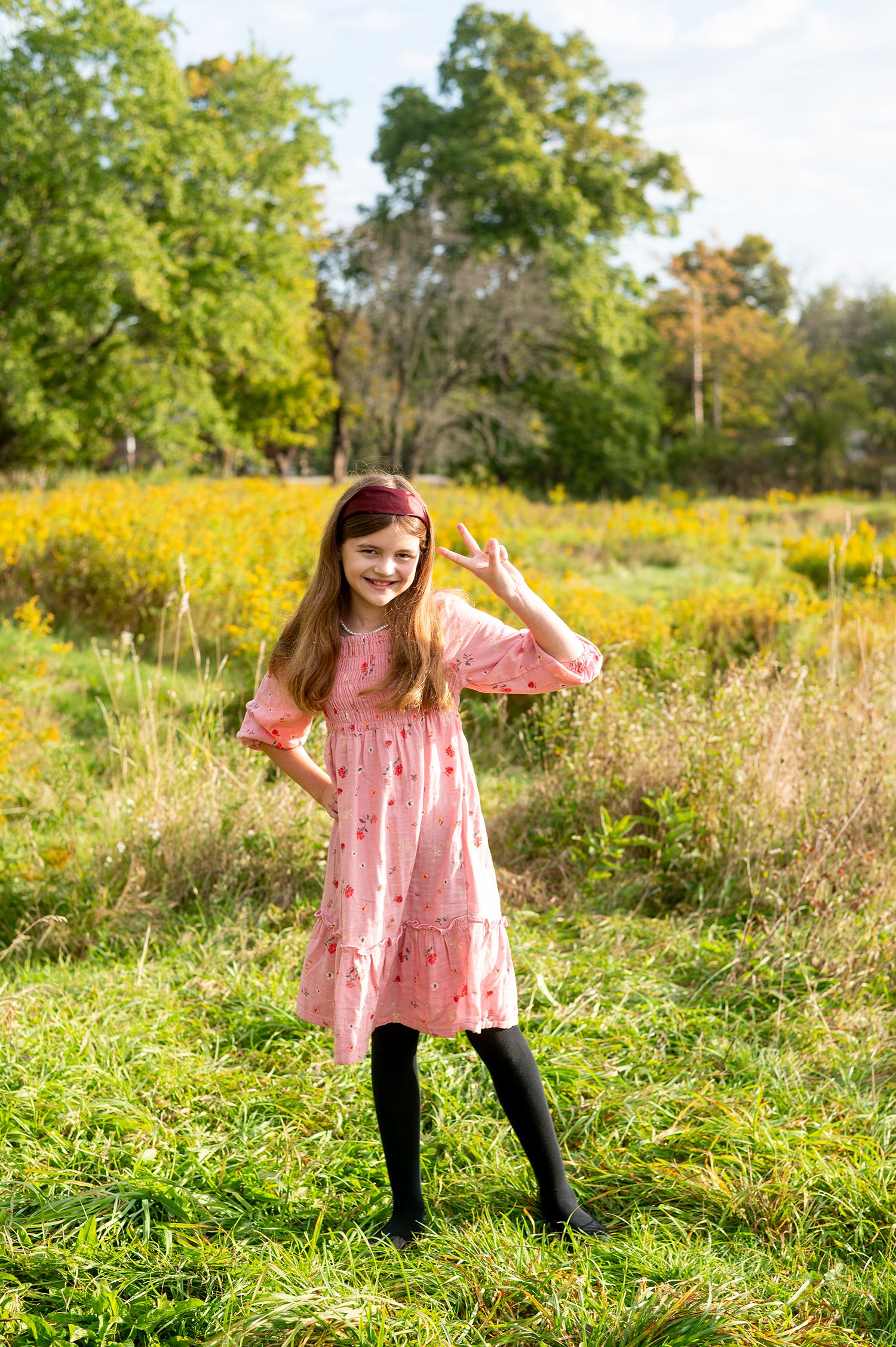 Young girl in pink dress and black boots waving in a sunny field with yellow wildflowers and green trees.