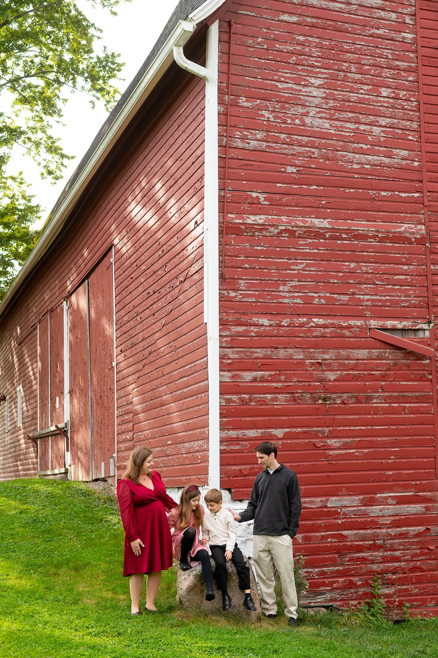 Family posing beside a rustic red barn with peeling paint on a grassy hillside.