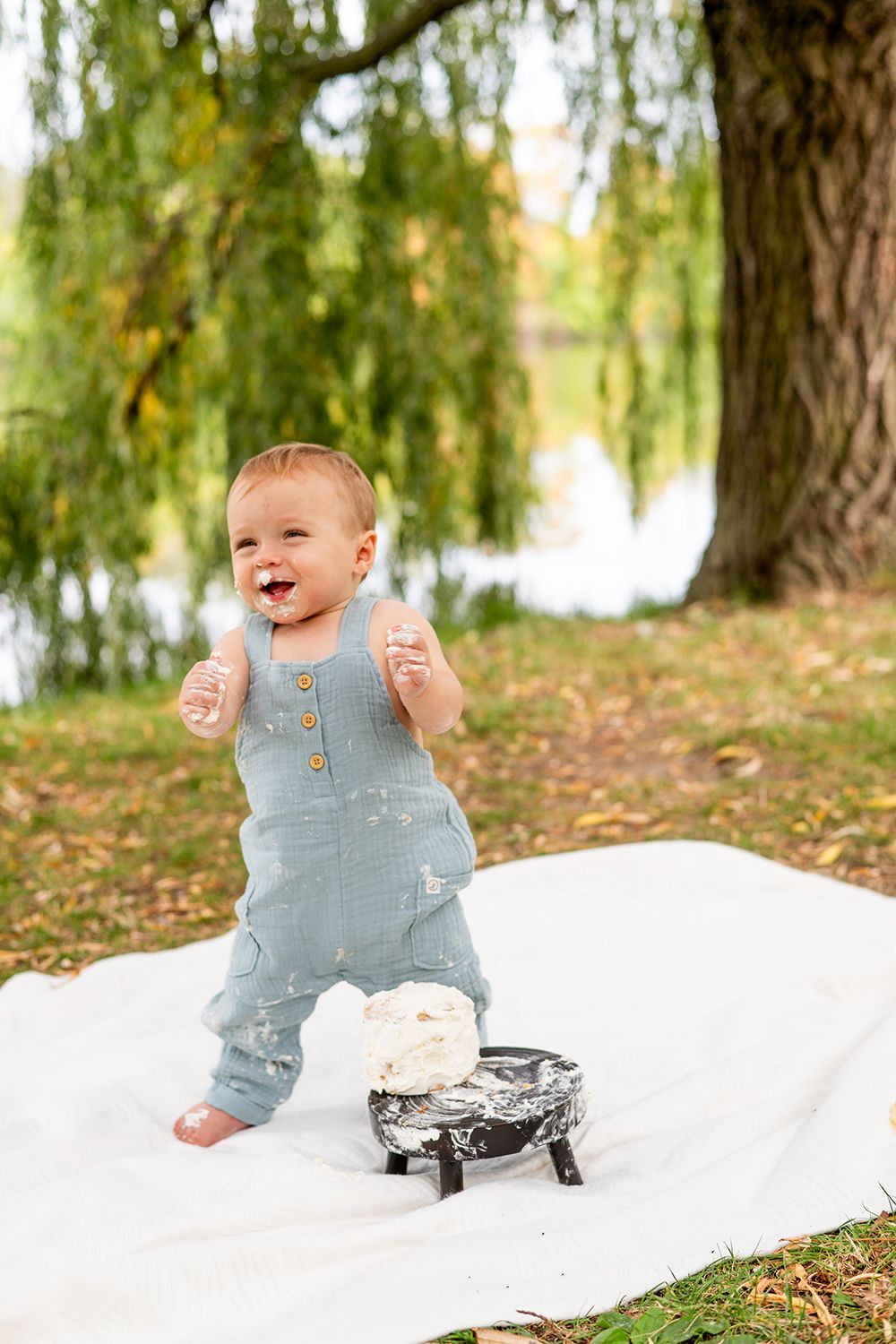 A toddler in light blue overalls stands joyfully on a white blanket under a willow tree by a pond.