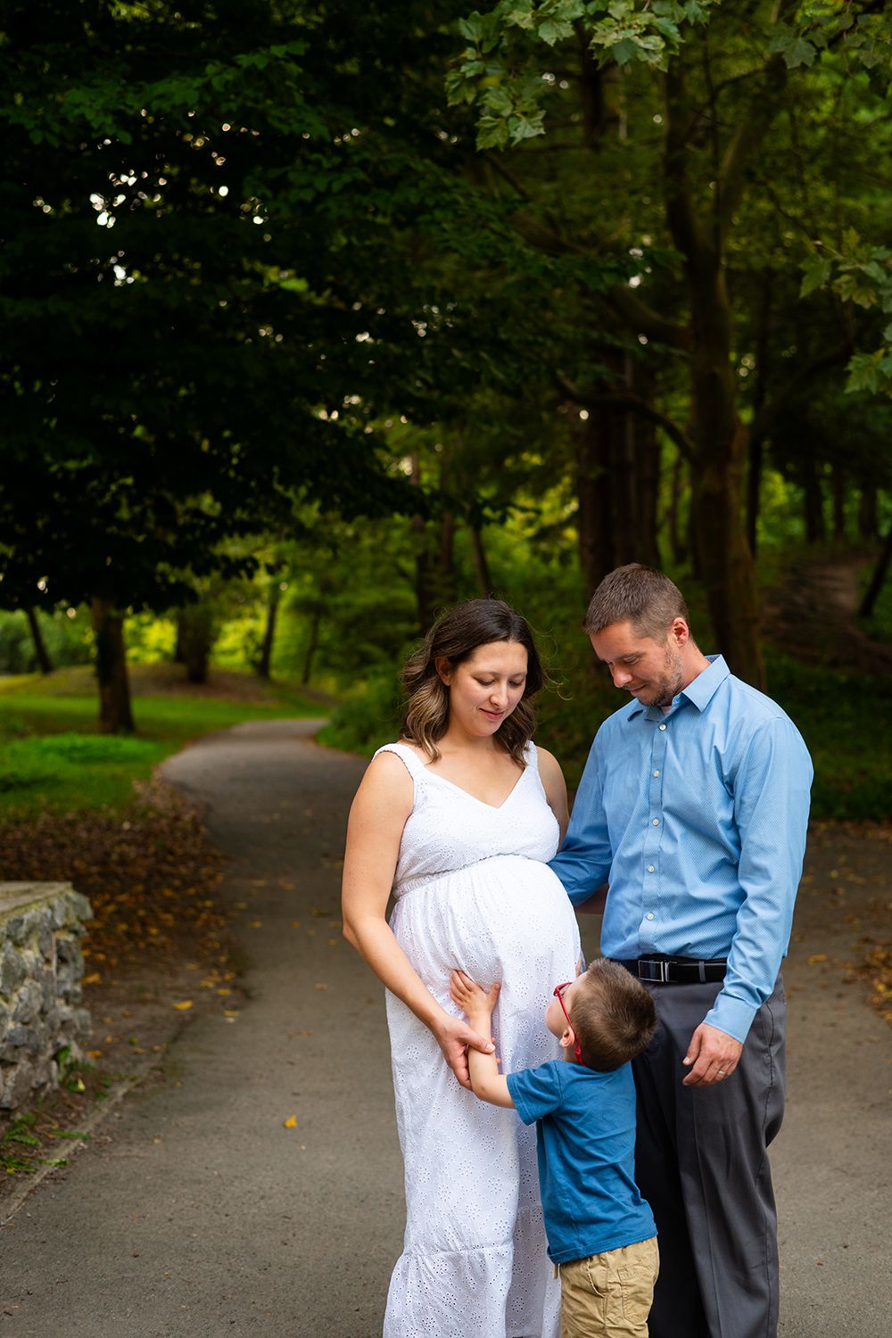 A family of three shares a tender moment on a tree-lined park path.
