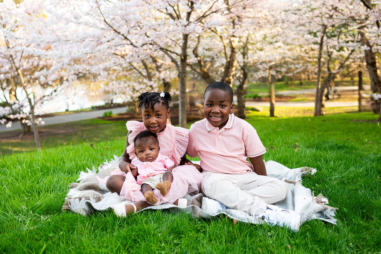 Family group wearing coordinated pink and white outfits relaxes together on green grass during spring cherry blossoms.