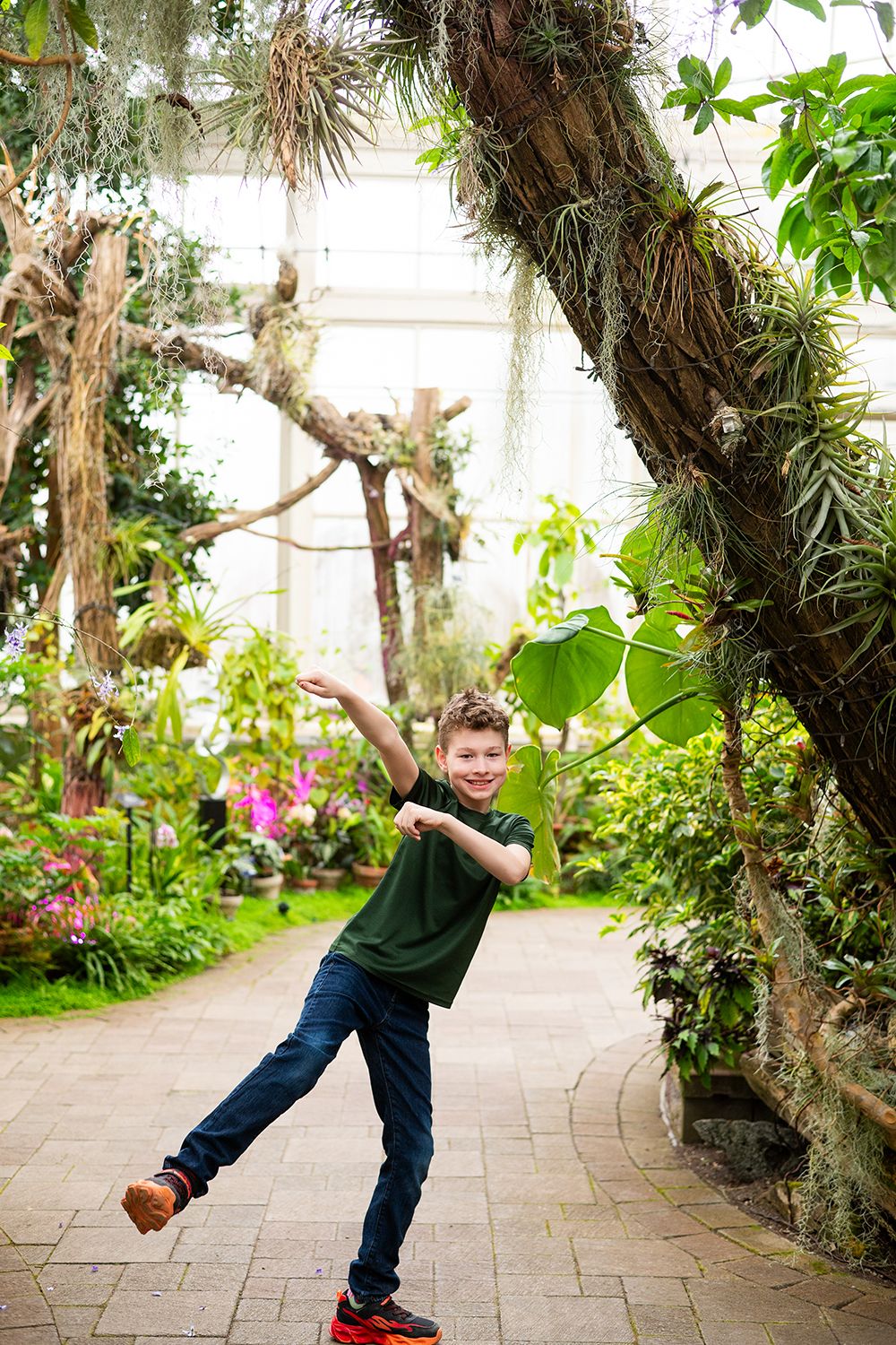 A playful child dances along a brick path in a lush greenhouse filled with tropical plants and flowers.