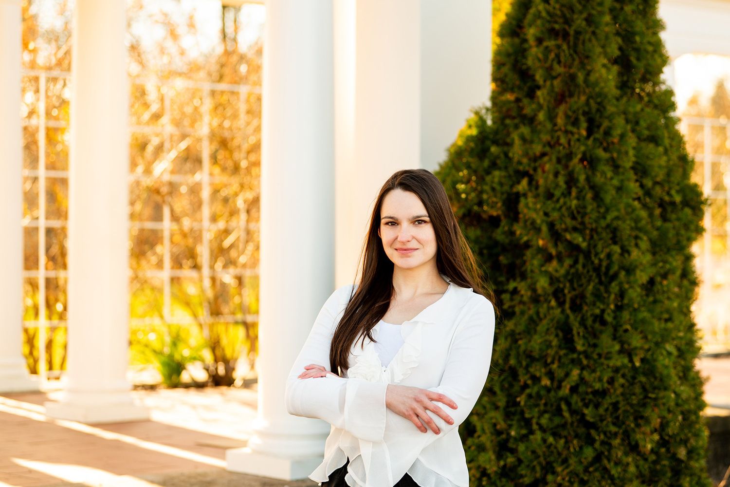 Business professional standing and smiling outdoors alongside tall evergreen bushes and white columns at sunset.
