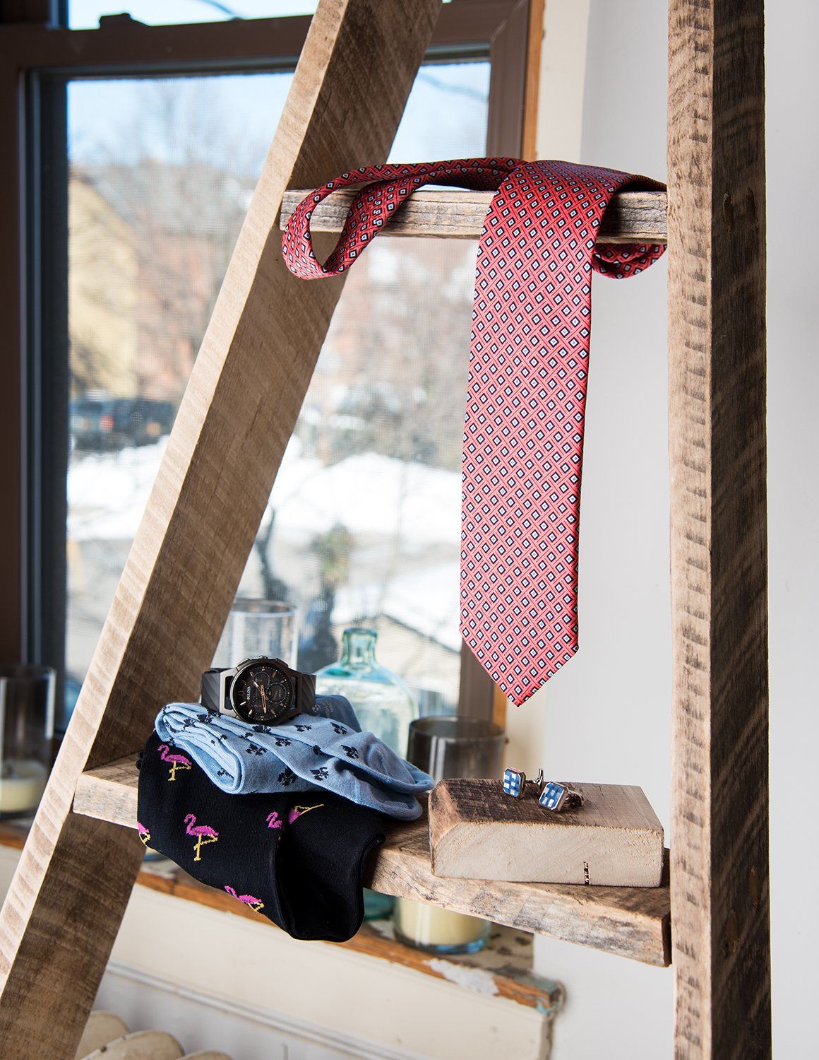 A pink patterned necktie hangs on a wooden ladder shelf with folded clothing and accessories near a window.