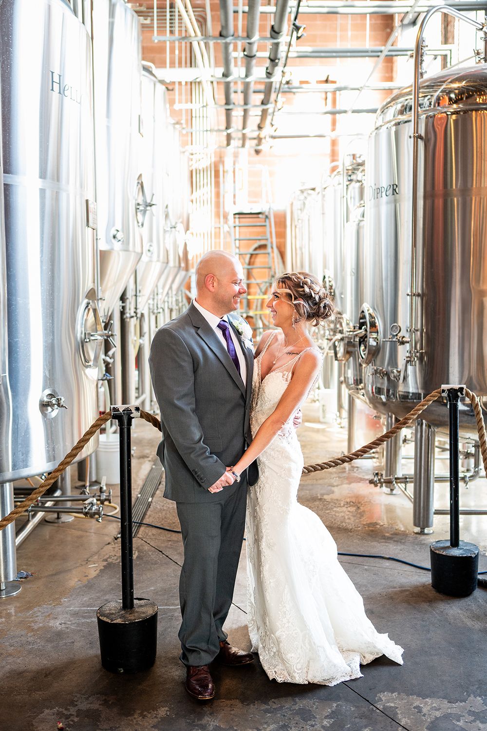 A couple embraces in a brewery setting surrounded by large metal fermentation tanks and industrial equipment.