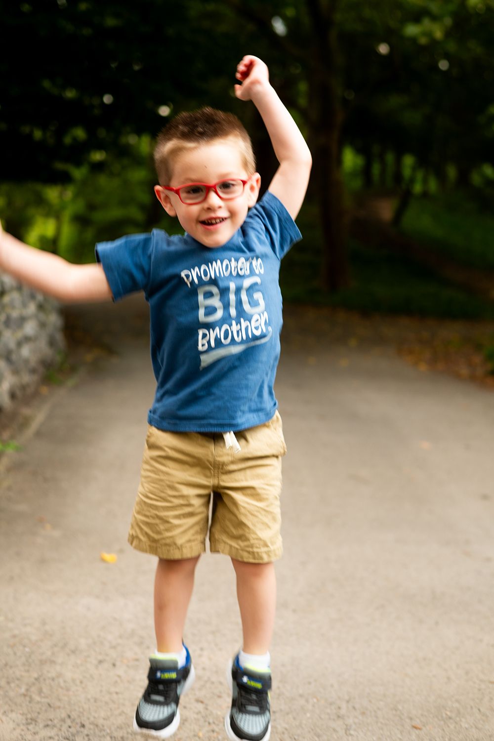 Child in a blue 'Big Brother' t-shirt and khaki shorts jumps excitedly on a wooded path.
