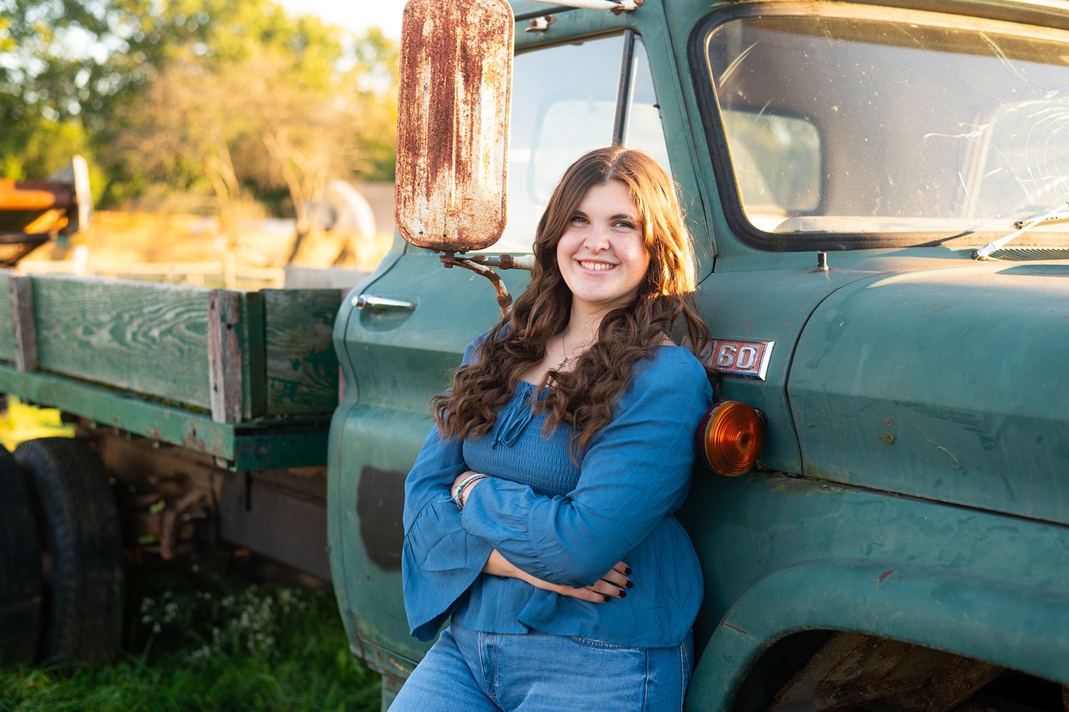 A person in denim outfit leans against a vintage green truck in warm afternoon light.