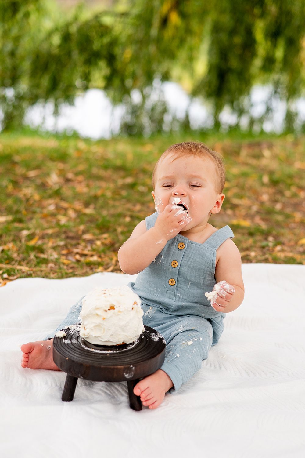 A toddler in denim overalls sits on a white blanket enjoying cake during an outdoor photoshoot.