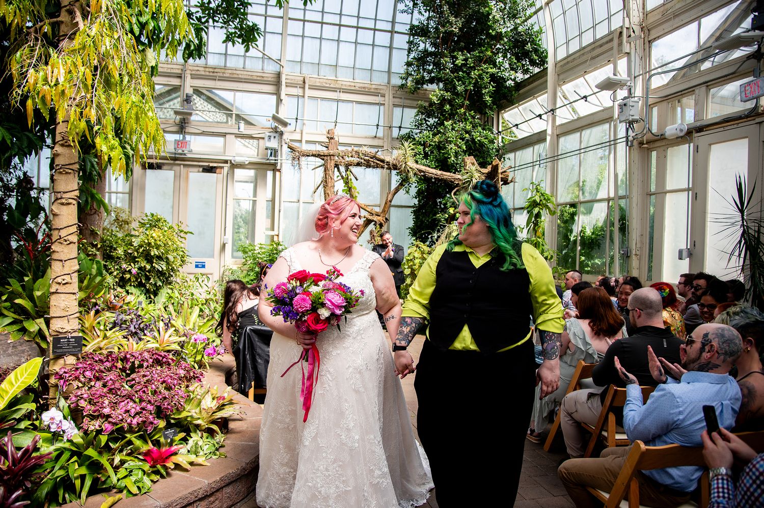 Two brides walk down the aisle in a lush greenhouse wedding, one in white lace with pink bouquet, other in black and green outfit.