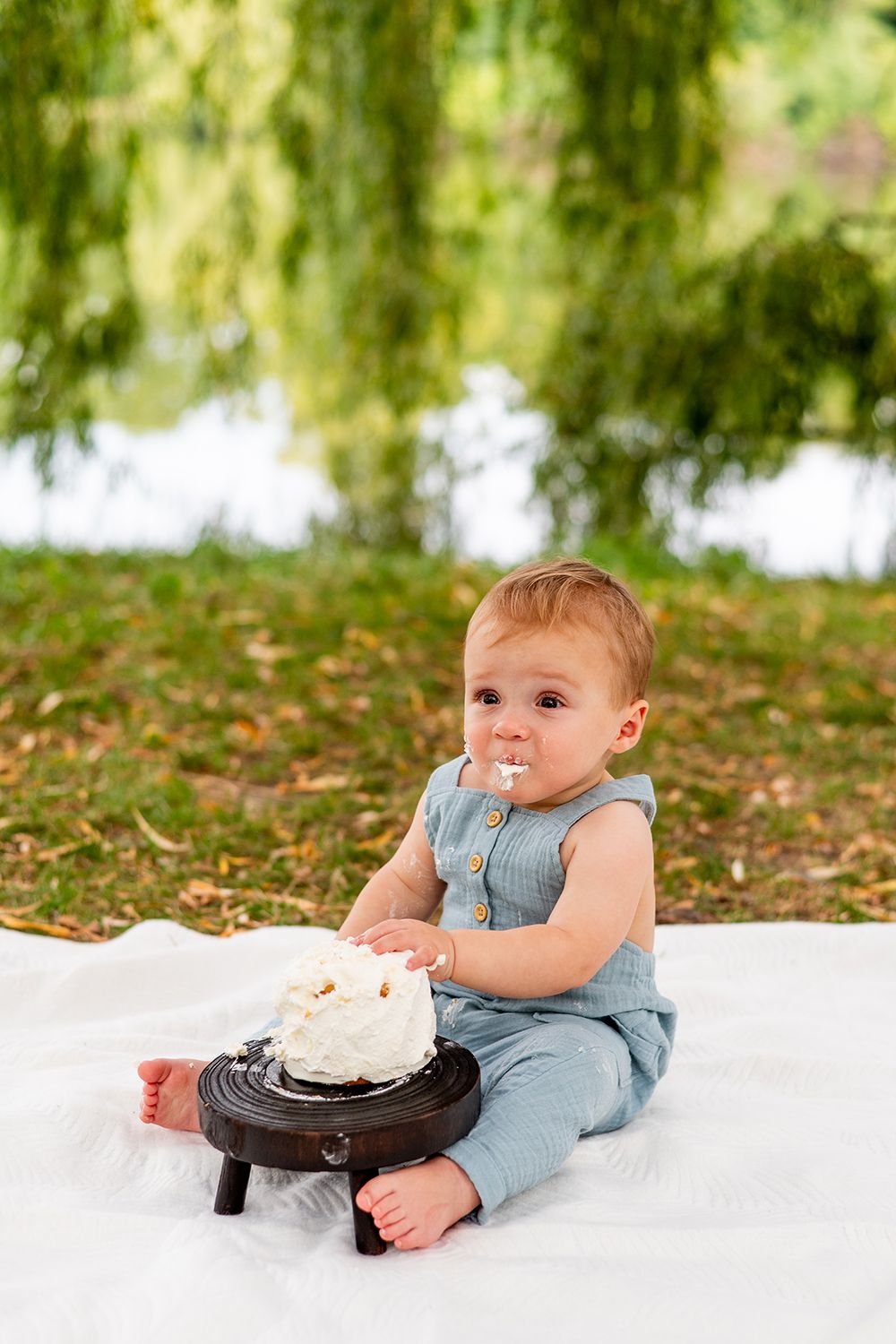 Small child in blue overalls sits on white blanket outdoors with cake during celebration.