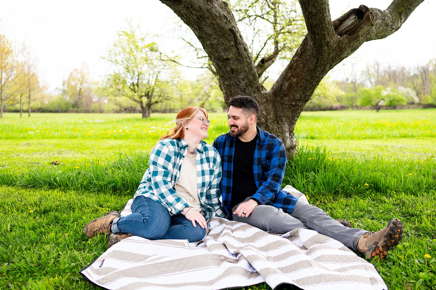 Couple shares intimate moments on picnic blanket under flowering tree in sunny spring meadow.