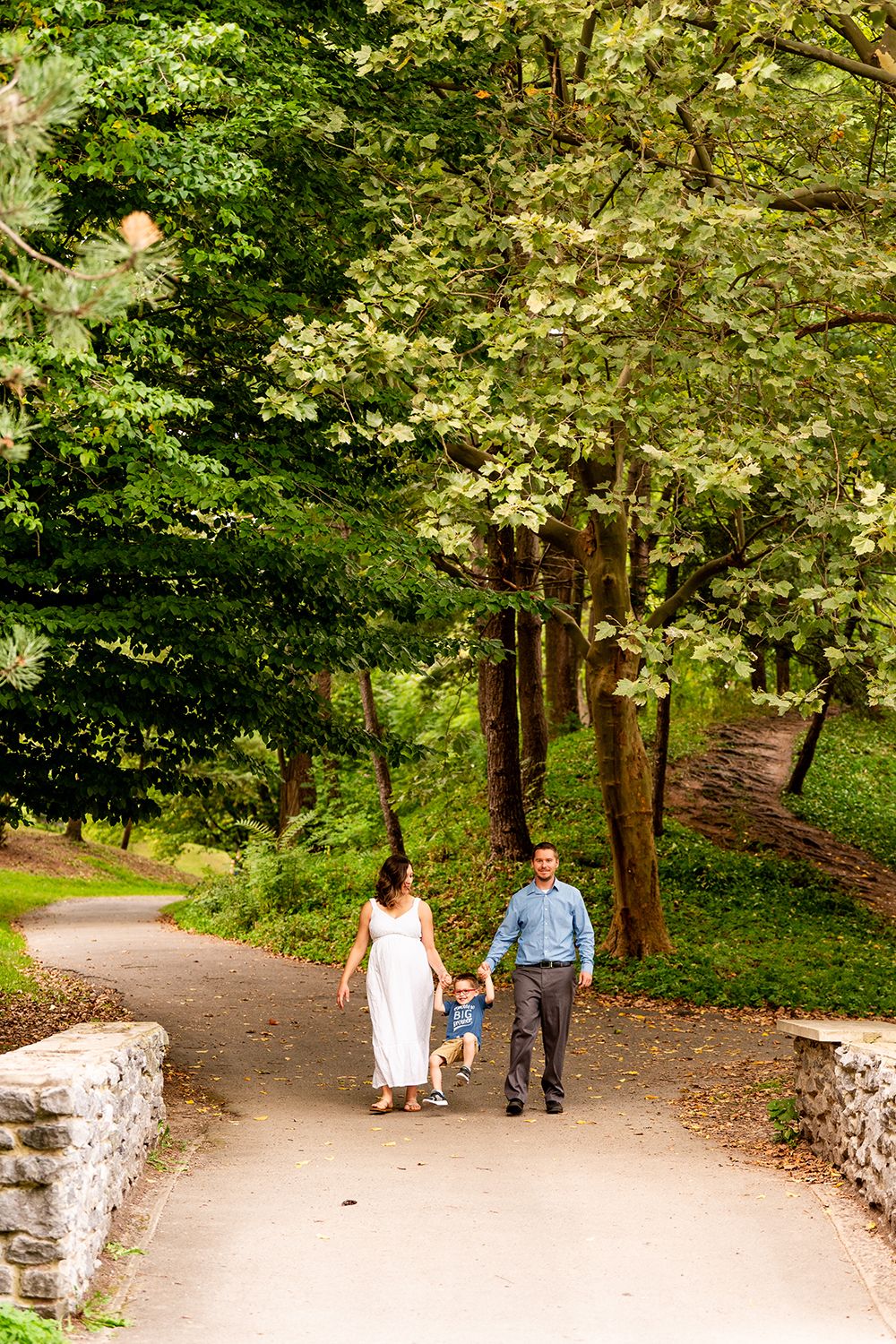 Couple strolls together on a winding park path surrounded by lush green trees and white stone walls.
