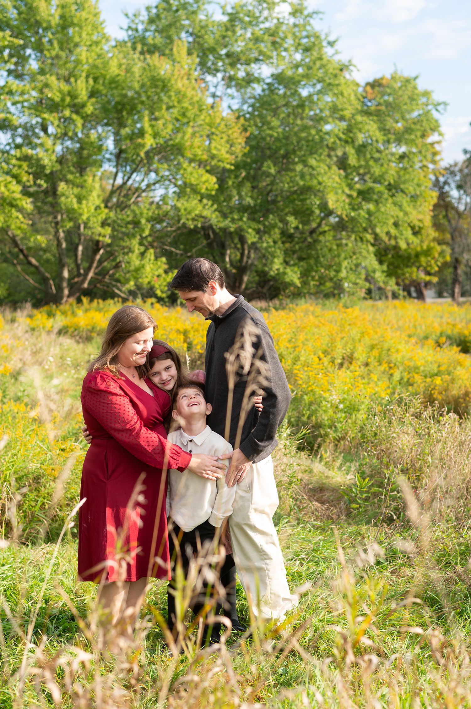 Family of four sharing a joyful moment in a sunny wildflower meadow with lush green trees in the background.