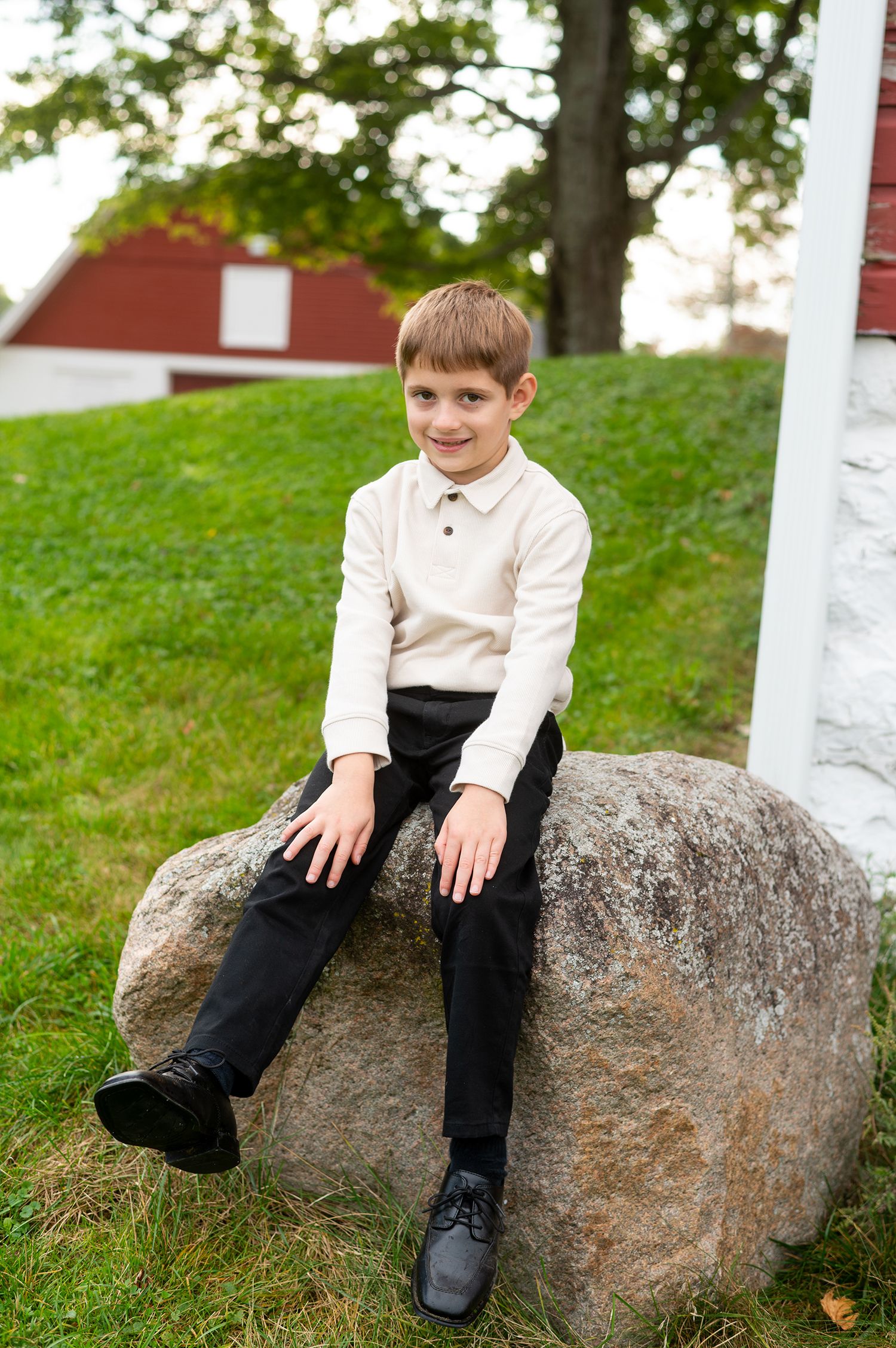 A smiling boy in a white sweater and black pants sits on a large rock outdoors near a red barn.