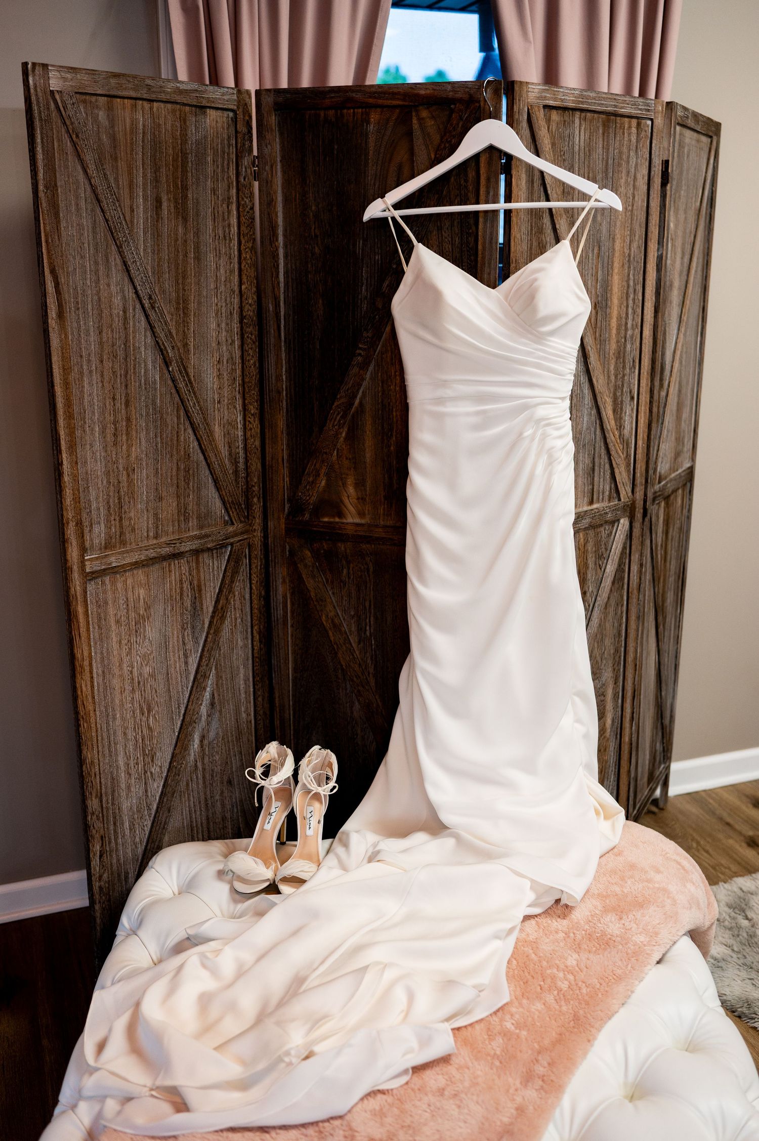 A white wedding dress hangs elegantly on a wooden hanger against a rustic barn door with delicate lace details and matching shoes.