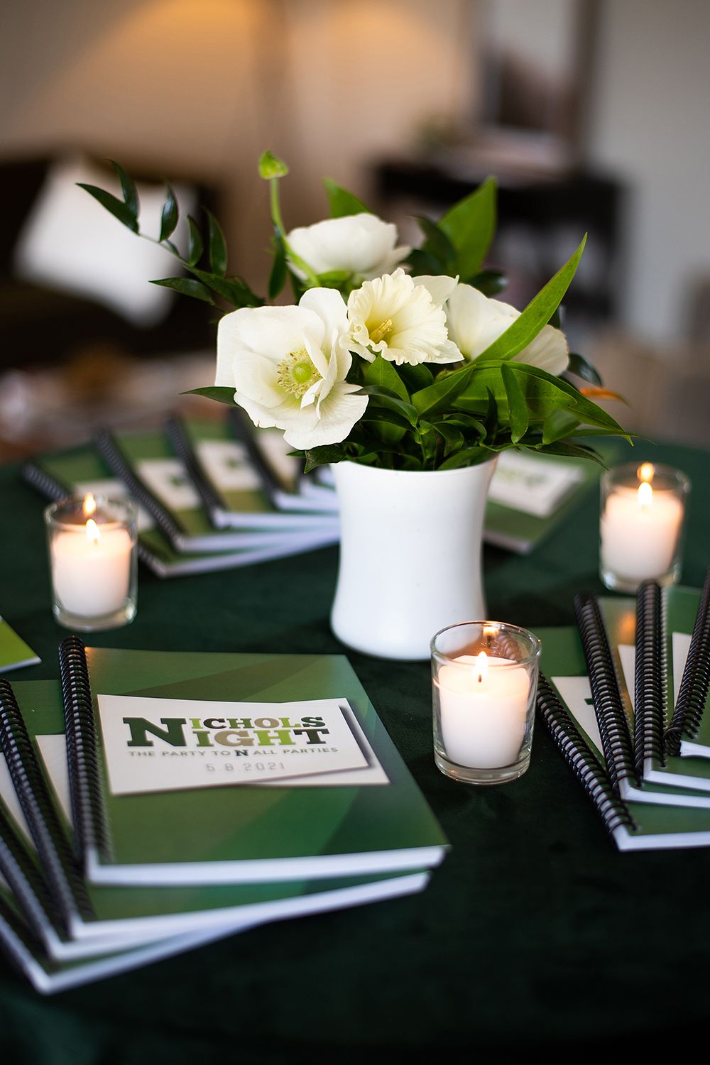 Table decor with white floral centerpiece, candles and black notebooks on dark green tablecloth for Movie Night event.