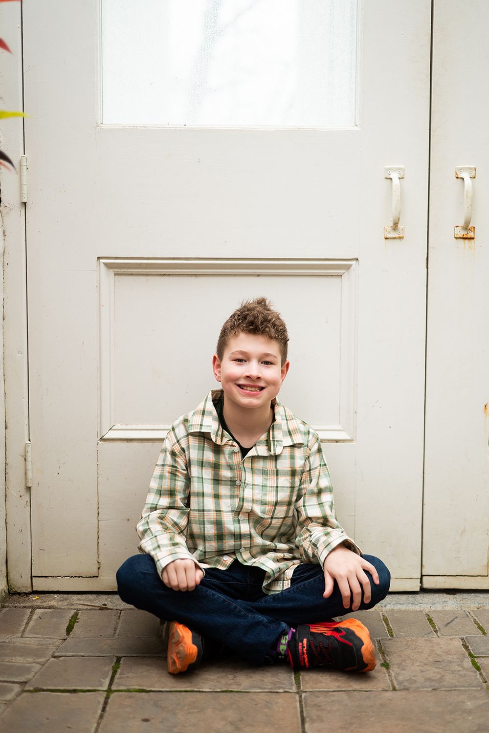 A young person in a plaid shirt and jeans sits cross-legged on a stone floor in front of a white door.