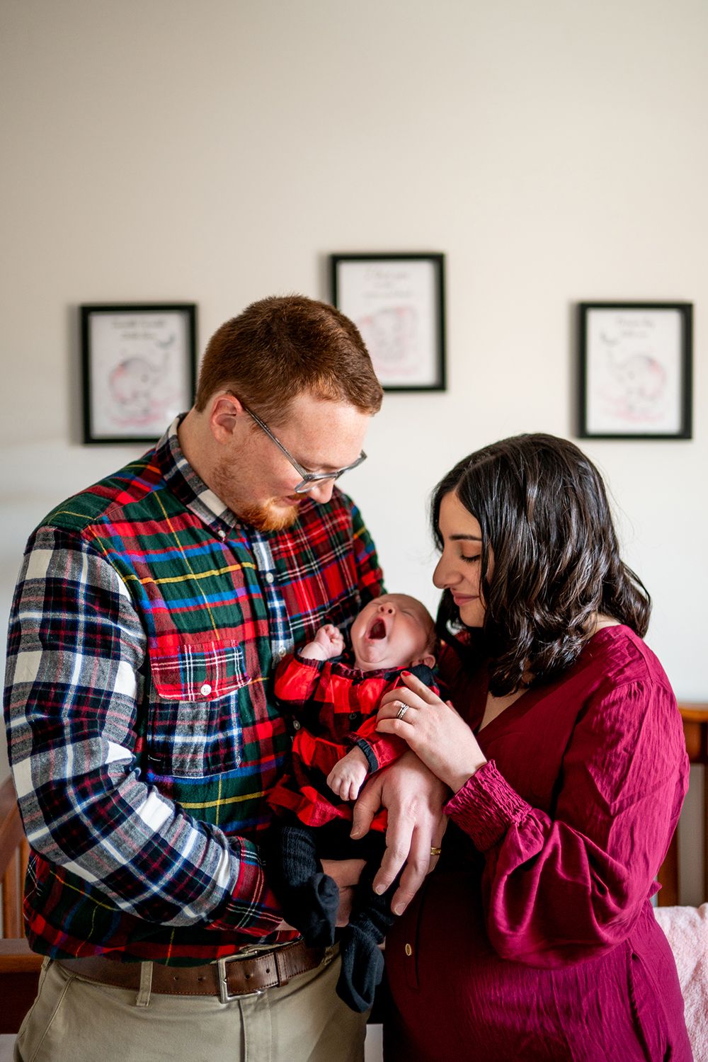 A couple in red and plaid outfits share a tender moment with their baby by a wall with framed artwork.