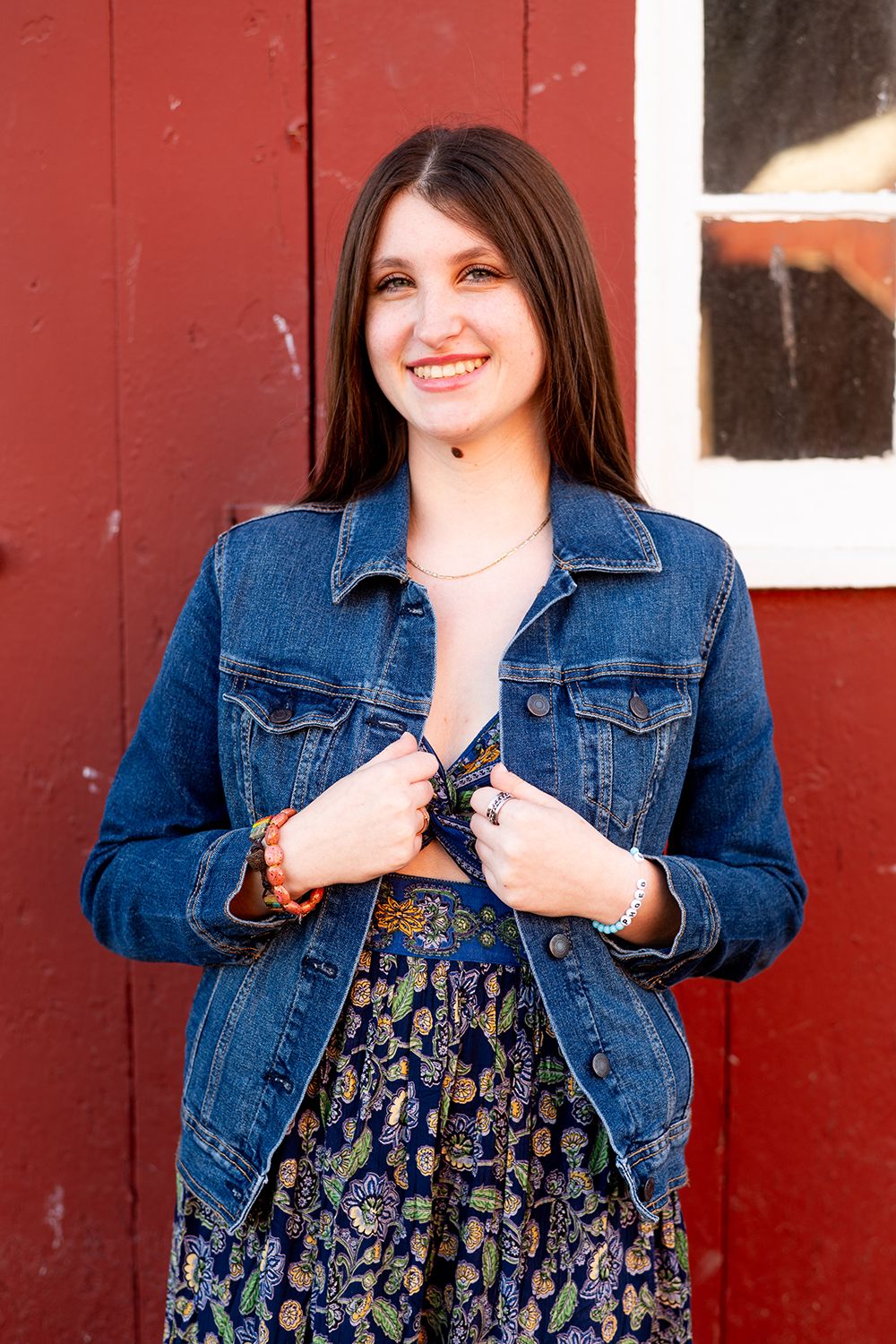 Someone wearing a denim jacket and floral dress poses against a red wall with a warm smile.