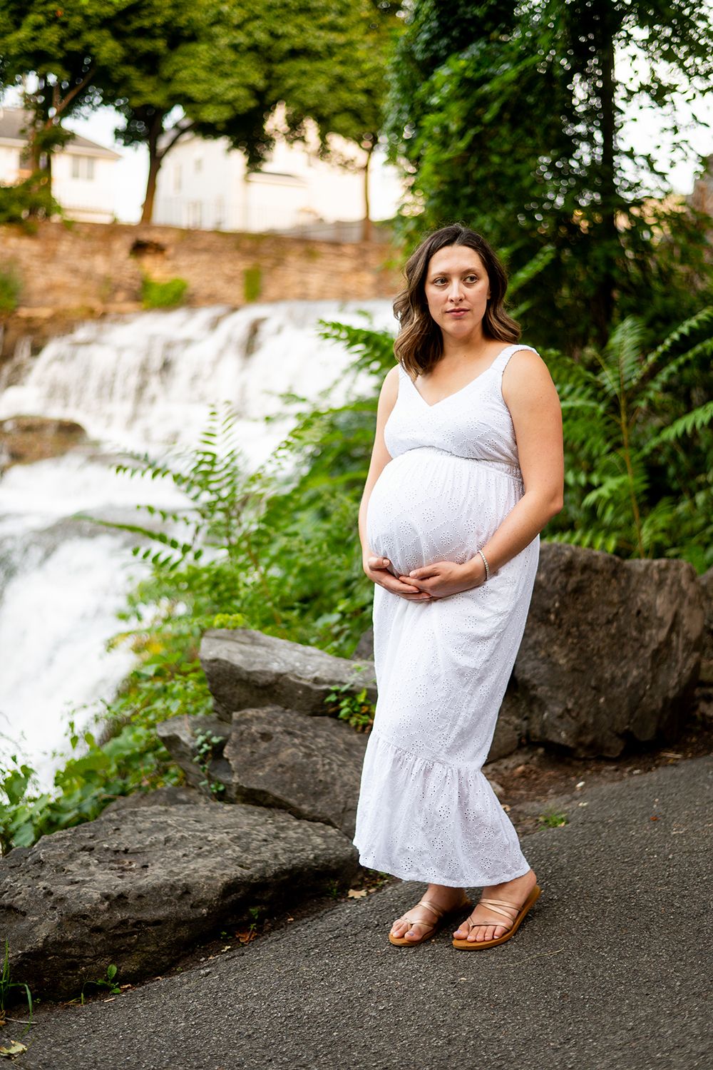 Woman in white maxi dress stands by scenic waterfall on nature path during maternity photo shoot.