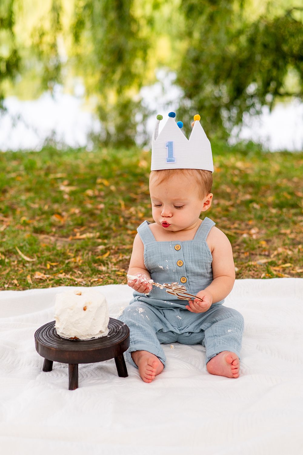 A small child in a paper crown and blue outfit sits on a blanket with a smash cake for their first birthday.