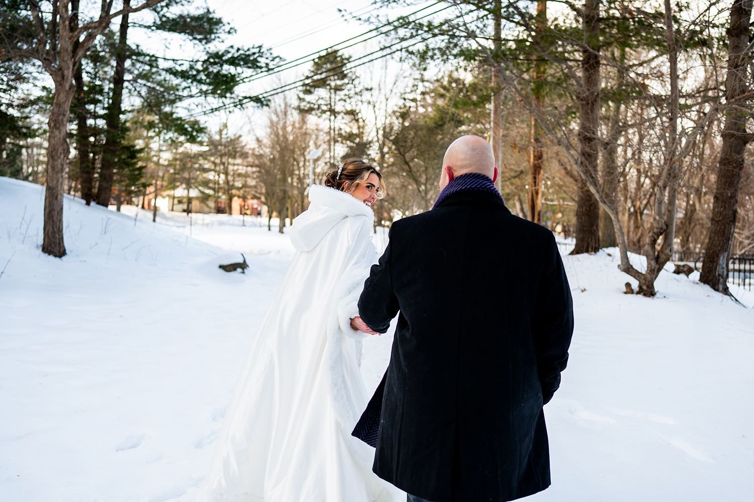 A couple walks together through a snowy winter forest during their wedding photoshoot.