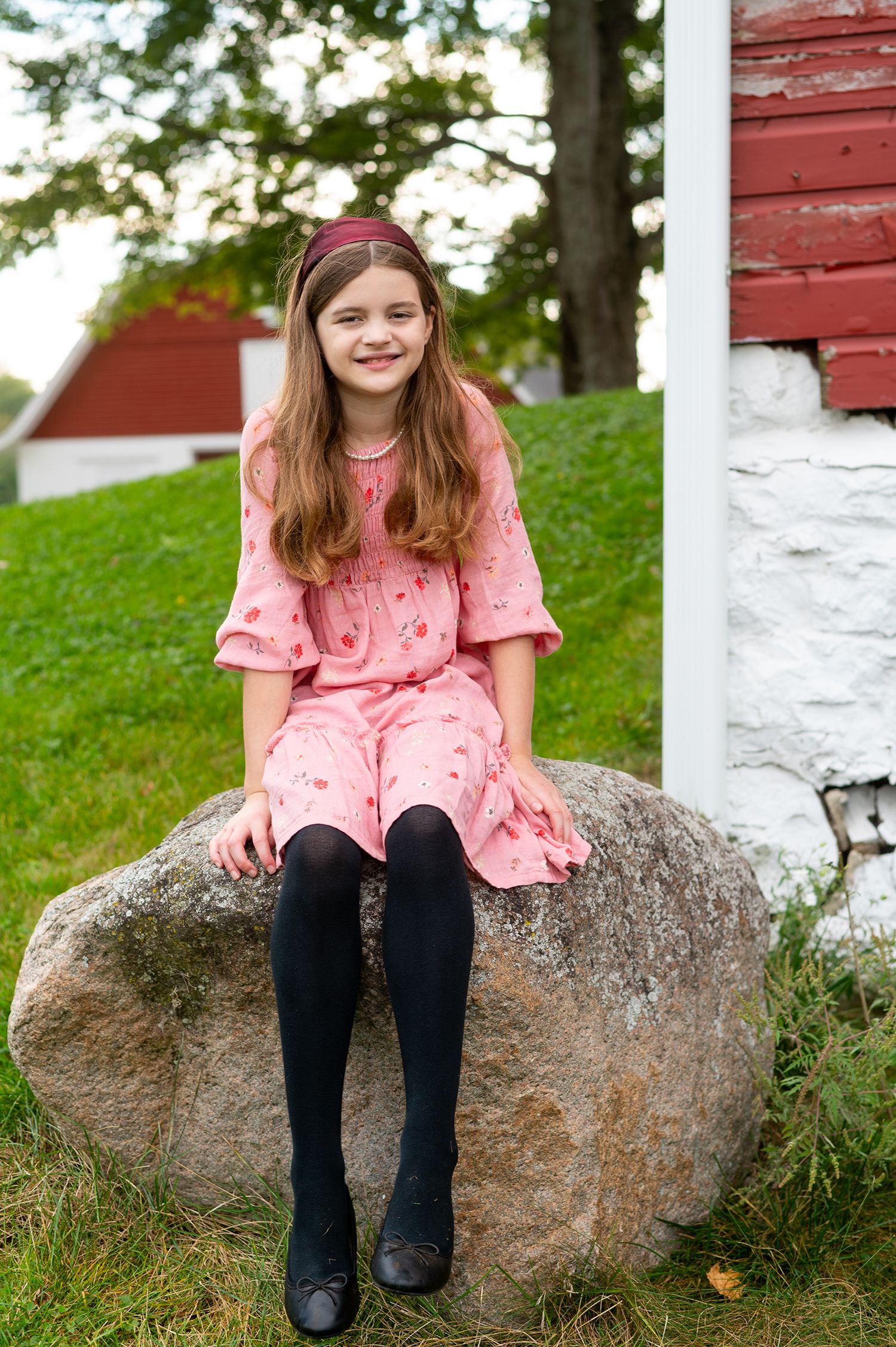 A young girl in a pink floral dress and black tights sits smiling on a large rock near a white and red barn.