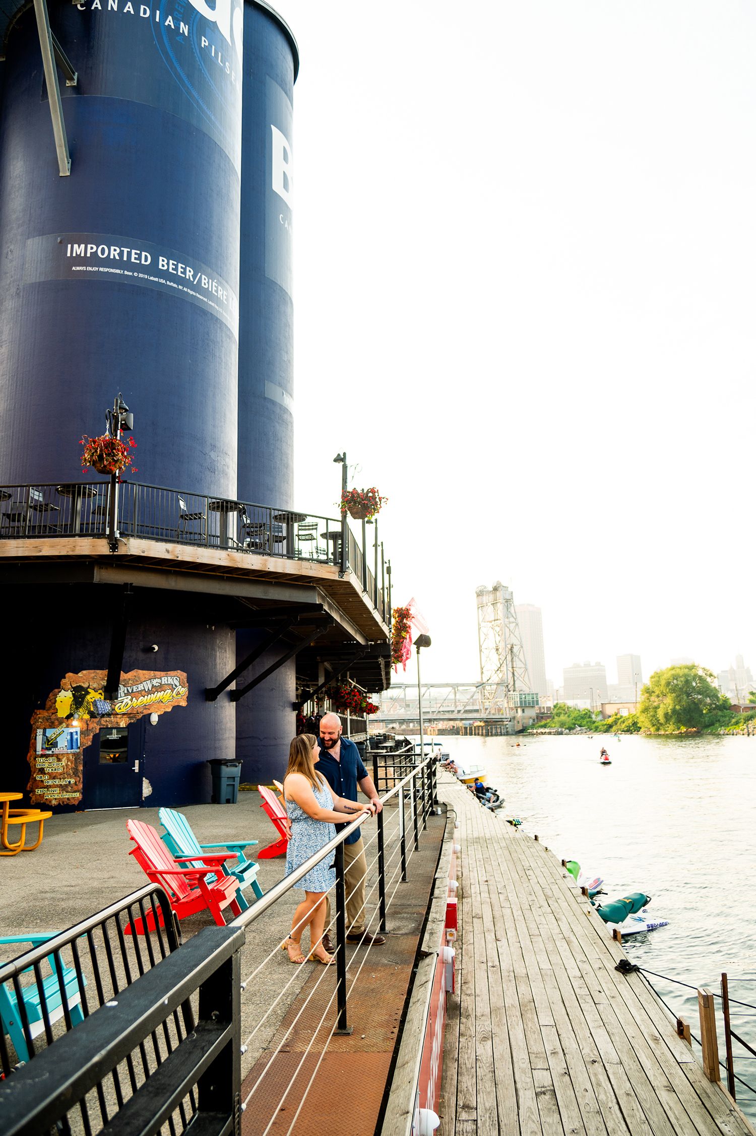 Couple enjoying waterfront views from a deck beside a blue cylindrical building with imported beer signage.