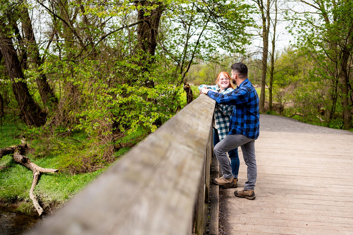 People sit and stand along a wooden boardwalk path winding through a lush green forest on a sunny day.