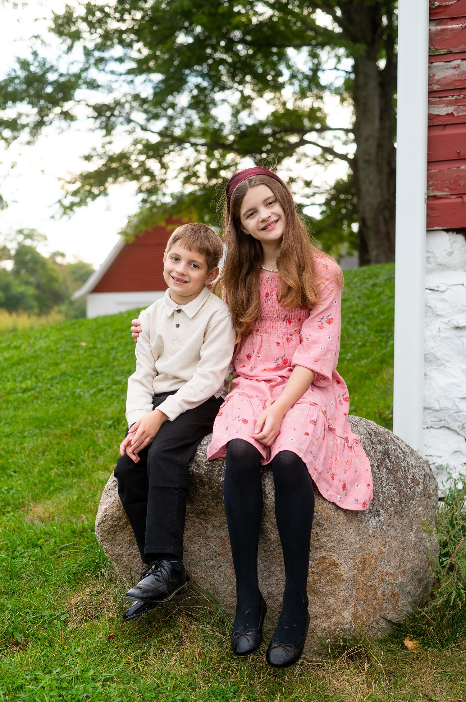 Two children, a boy and girl, sit smiling on a large rock outdoors near a red barn.