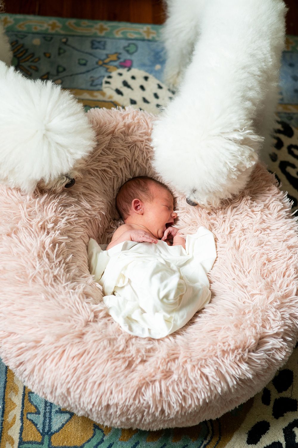 A sleeping newborn in a white outfit rests peacefully on a plush pink fuzzy cushion surrounded by white pillows.