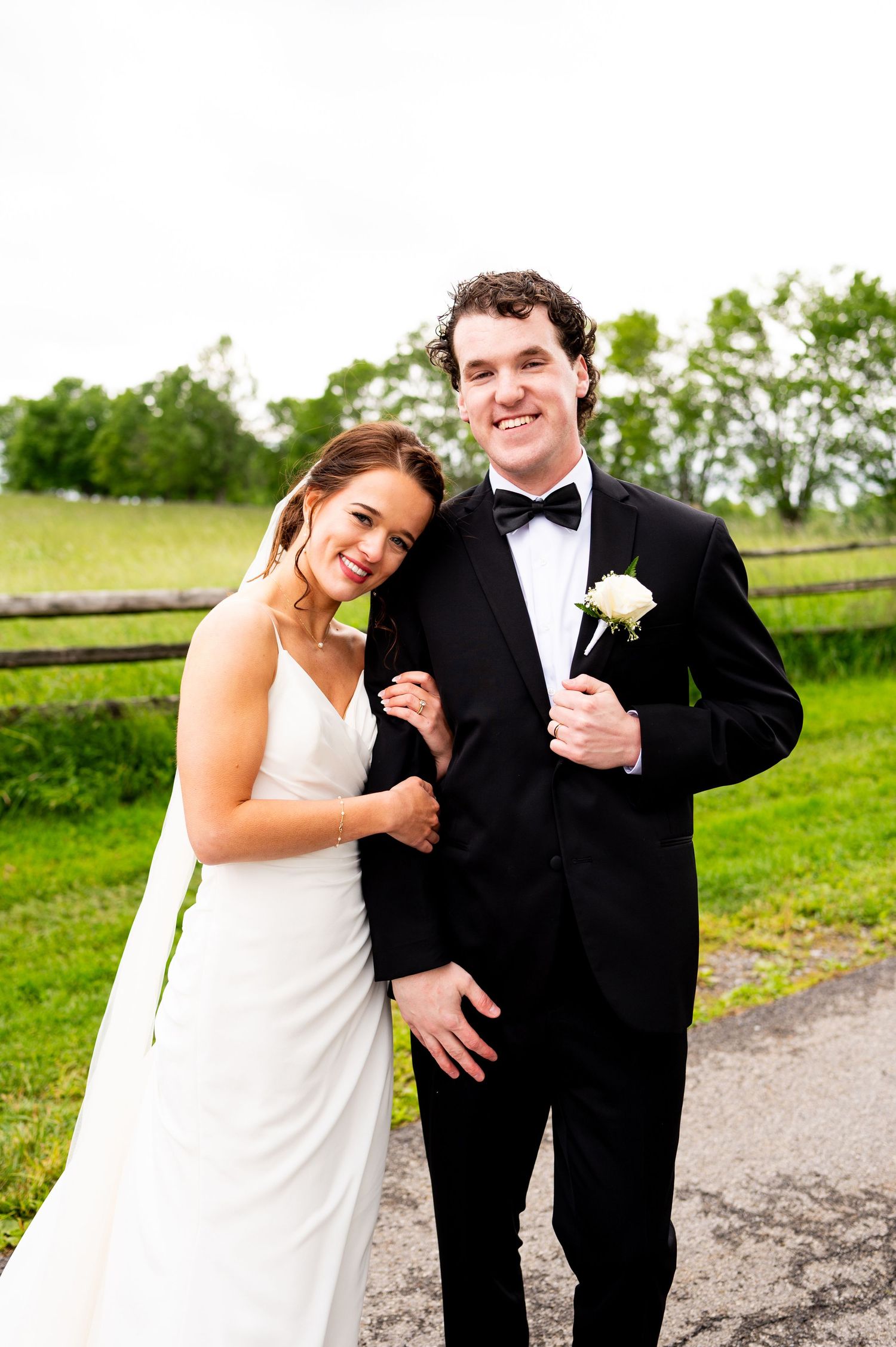 A couple in wedding attire shares joyful candid moments together on a country path with green fields in background.