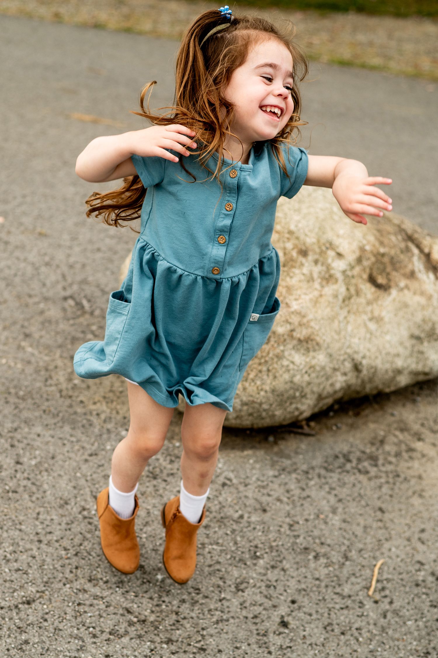 A happy little girl in a teal dress and brown boots jumping and spinning outdoors.