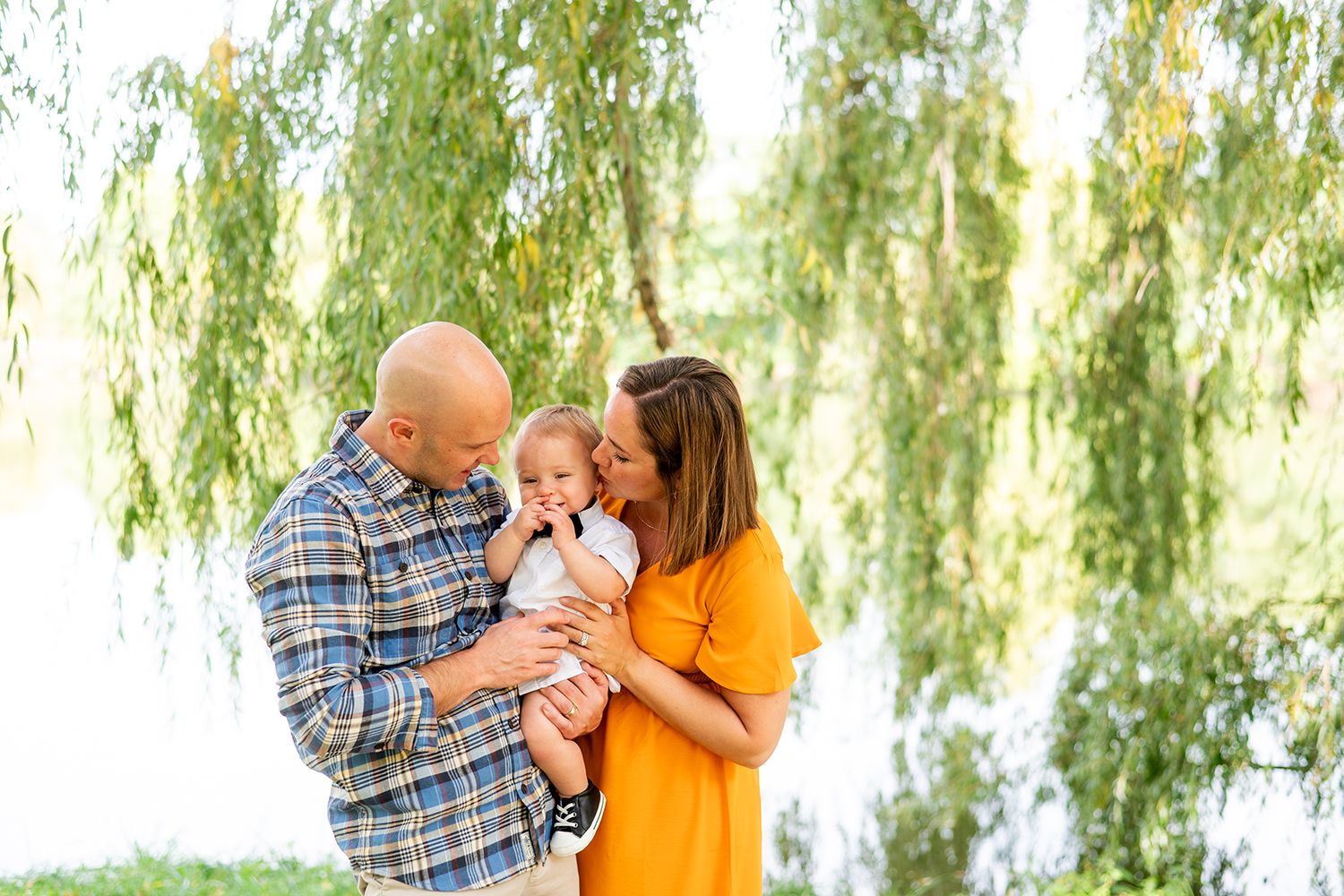 Family shares tender moment together outdoors under willow trees beside a lake on sunny day.