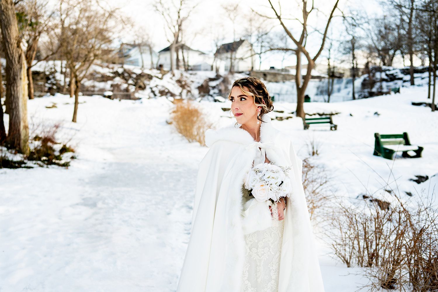 A person in a white winter wedding dress twirls joyfully in the snow with houses and trees behind them.