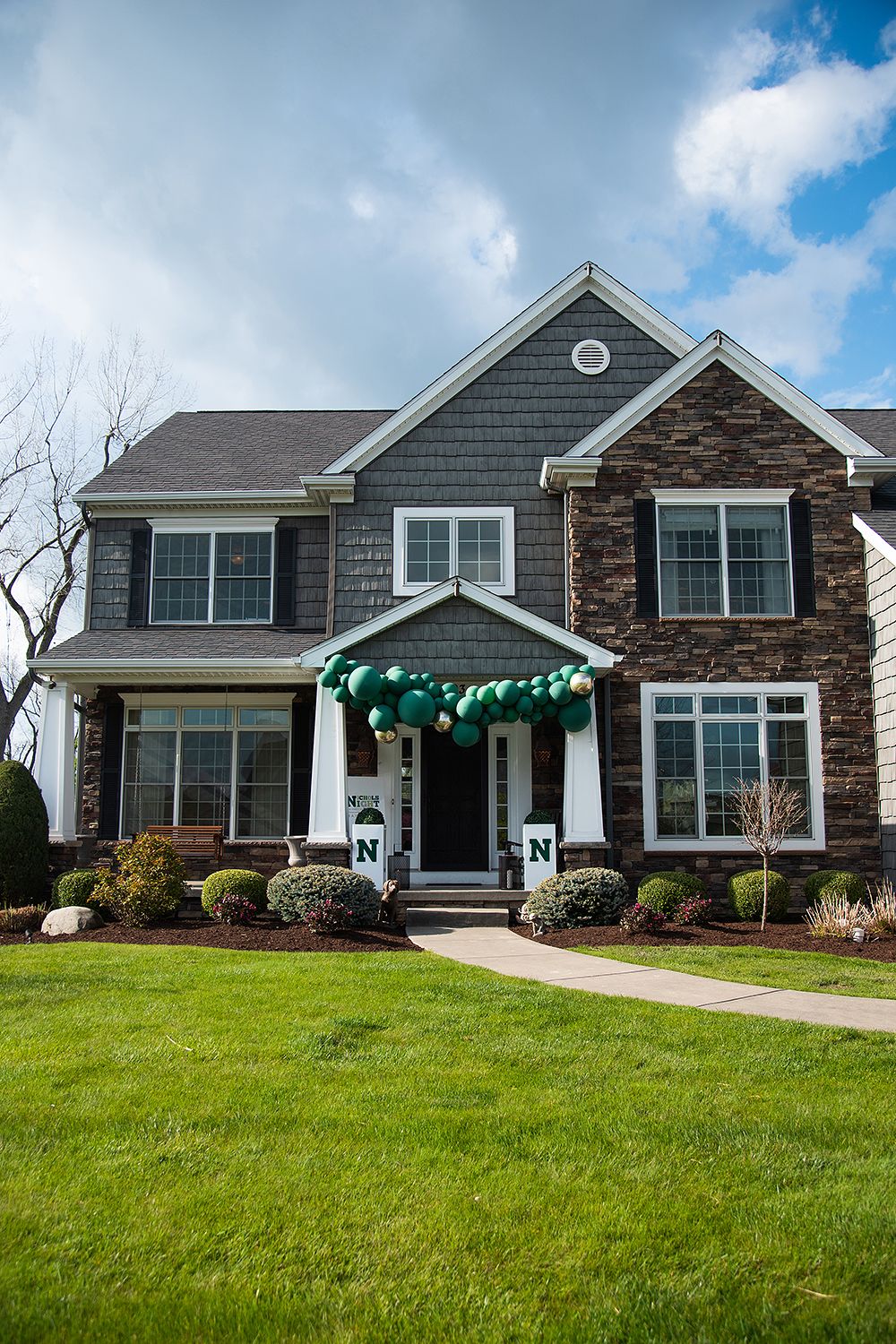 Modern craftsman-style house with stone accents and gray siding features a well-manicured lawn under blue skies.