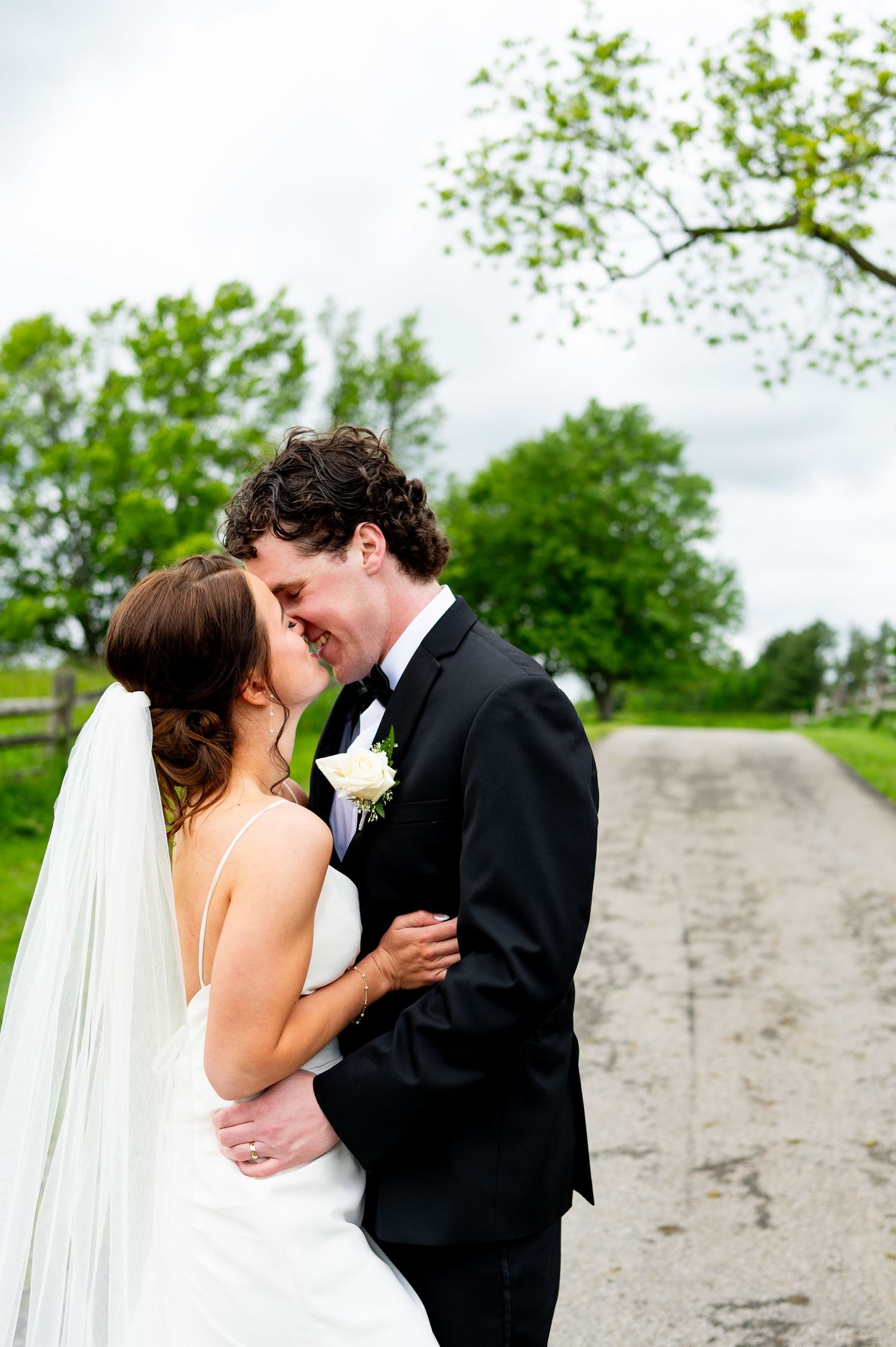 Newlyweds embrace in a passionate kiss along a stone wall with lush greenery surrounding them.