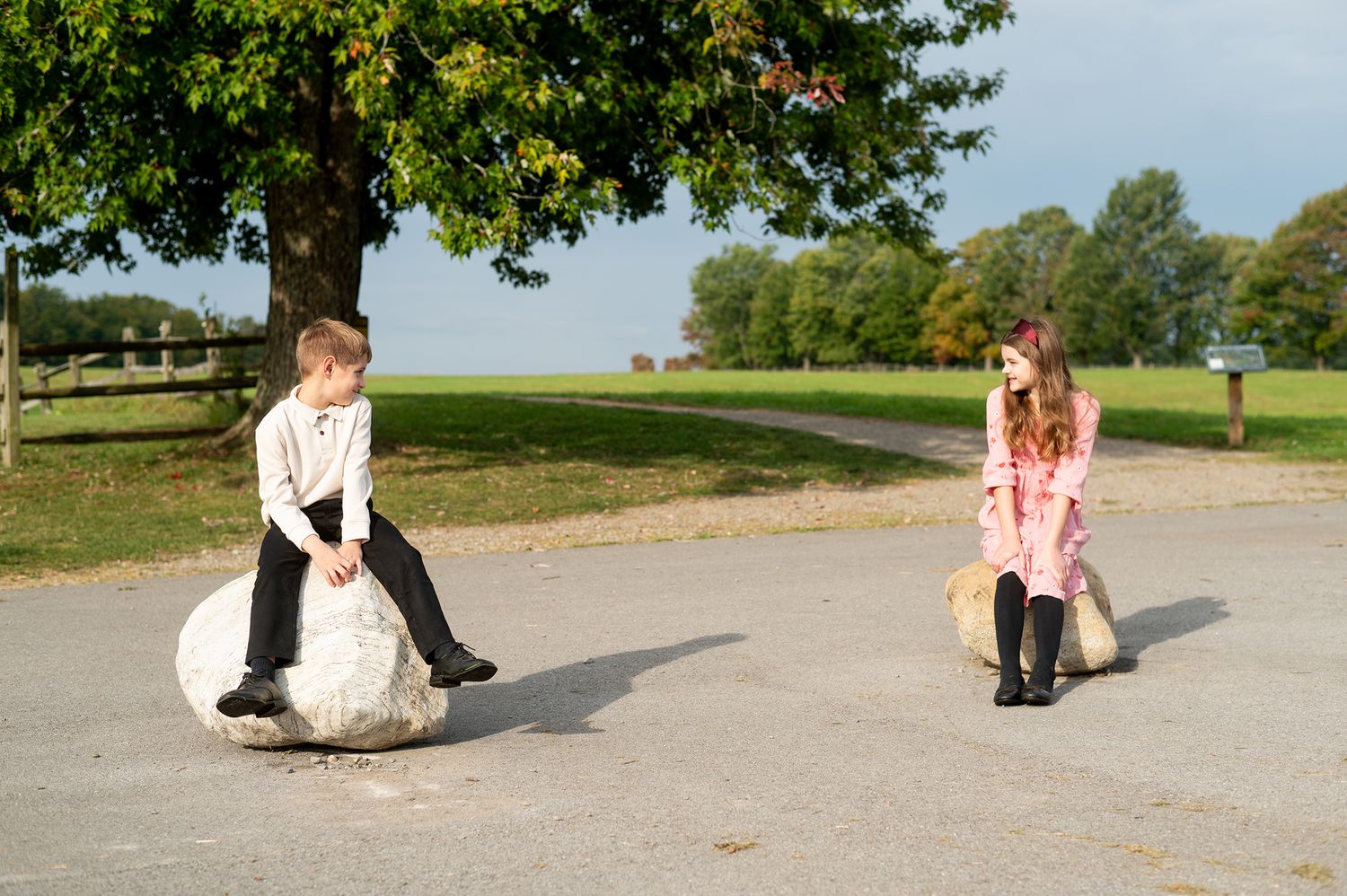 Two children sitting on large rocks in a park, a boy in white shirt and a girl in pink dress, facing each other.